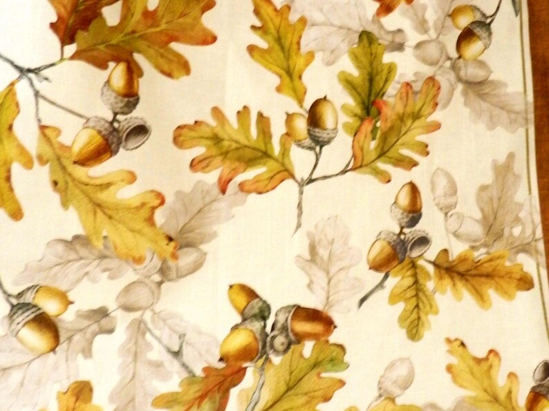 K&ouml;nnte beinhalten: Ein Textilstoff mit einem sich wiederholenden Muster aus Eichenbl&auml;ttern und Eicheln. Das Design umfasst leuchtend gelbe, orangefarbene und gr&uuml;ne Bl&auml;tter neben graut&ouml;nigen Bl&auml;ttern sowie goldbraune Eicheln auf einem cremefarbenen Hintergrund.