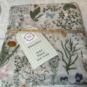 K&ouml;nnte beinhalten: Ein gefaltetes Baumwoll-K&uuml;chenhandtuch mit botanischem Druck aus gr&uuml;nen Kr&auml;utern, Wildblumen und blauen Schmetterlingen auf wei&szlig;em Grund. Das Handtuch ist mit Bindfaden verschn&uuml;rt und hat ein hellgr&uuml;nes Vichy-Karo-Etikett mit der Aufschrift Antique Marie.