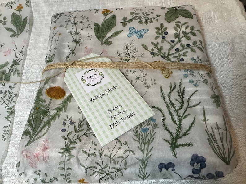 K&ouml;nnte beinhalten: Ein gefaltetes Baumwoll-K&uuml;chenhandtuch mit botanischem Druck aus gr&uuml;nen Kr&auml;utern, Wildblumen und blauen Schmetterlingen auf wei&szlig;em Grund. Das Handtuch ist mit Bindfaden verschn&uuml;rt und hat ein hellgr&uuml;nes Vichy-Karo-Etikett mit der Aufschrift Antique Marie.