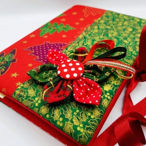 May include: A red and green Christmas-themed fabric journal with a red ribbon and a green and red fabric flower with a white polka dot button.