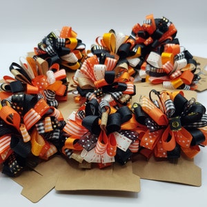 Puede incluir: Un conjunto de ocho lazos para el pelo con temática de Halloween en cinta negra, naranja y blanca. Los lazos están hechos con múltiples capas de cinta y tienen una variedad de patrones, que incluyen lunares, rayas y cuadros.