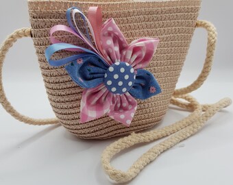 May include: A small beige straw bag with a flower-shaped embellishment. The flower is made of pink and blue fabric with white polka dots.