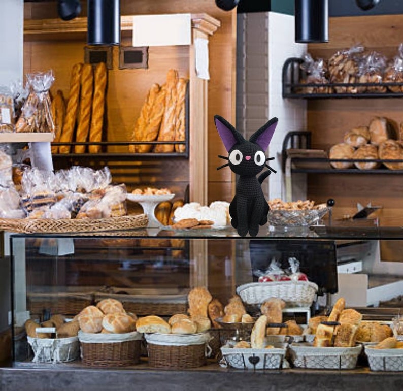 Puede incluir: Una figurita de gato de crochet negro con orejas moradas y ojos grandes se sienta en un mostrador frente a una vitrina de panader&iacute;a llena de varios panes y pasteles.