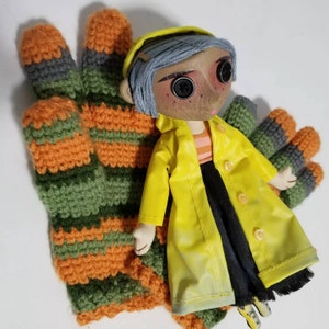 Puede incluir: Una muñeca hecha a mano que lleva un impermeable amarillo y una falda negra. La muñeca tiene el pelo azul y grandes ojos negros. La muñeca está sentada sobre un par de guantes de punto de rayas verdes y naranjas.