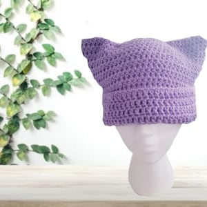 Può includere: Un berretto di lana color lilla con orecchie da gatto su una testa di manichino bianca.