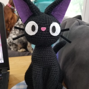 Puede incluir: Un gato negro de crochet con orejas moradas y ojos blancos. El gato est&aacute; sentado sobre una superficie a cuadros azules y blancos.