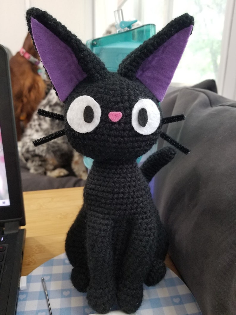 Puede incluir: Un gato negro de crochet con orejas moradas y ojos blancos. El gato est&aacute; sentado sobre una superficie a cuadros azules y blancos.