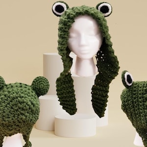May include: A green crocheted frog hat with large, round eyes and ear details. The hat is designed to cover the head and neck, with long, dangling flaps. The hat is displayed on a mannequin head, showcasing the whimsical design.