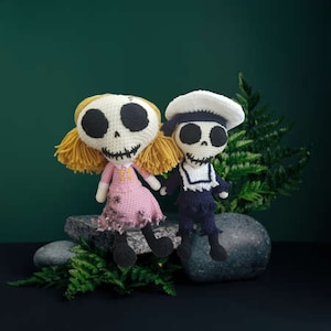 May include: Two handmade crochet dolls with skeleton faces. One doll wears a pink dress and has yellow hair, the other wears a sailor outfit and a white hat. Both dolls have black eyes and stitched smiles.