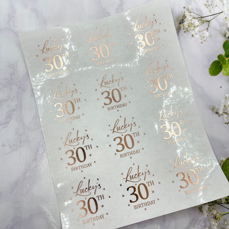May include: A sheet of clear stickers with rose gold foil lettering. The stickers say "Lucky's 30th Birthday".