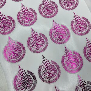 May include: A collection of glossy, circular stickers with a metallic pink finish. Each sticker features ornate calligraphy in a darker pink hue, arranged in a stylized teardrop shape. The stickers are arranged on a white surface.