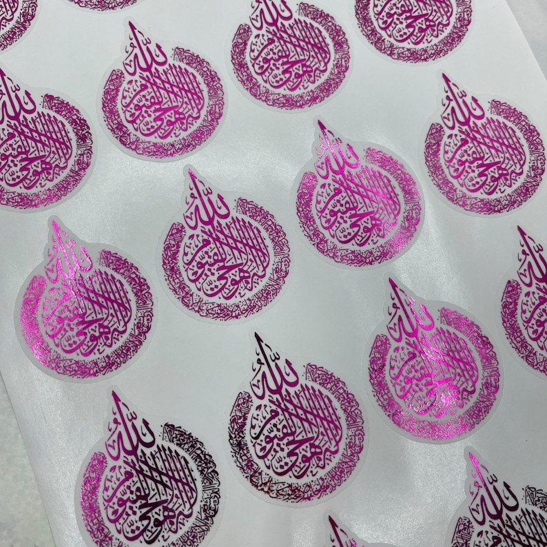 May include: A collection of glossy, circular stickers with a metallic pink finish. Each sticker features ornate calligraphy in a darker pink hue, arranged in a stylized teardrop shape. The stickers are arranged on a white surface.