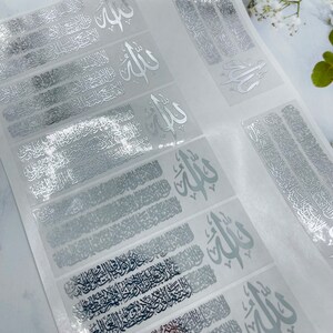 May include: A sheet of adhesive decals featuring silver metallic Arabic calligraphy. The design includes the word Allah in a prominent script, accompanied by intricate, repeating lines of detailed Arabic text arranged in horizontal rows.