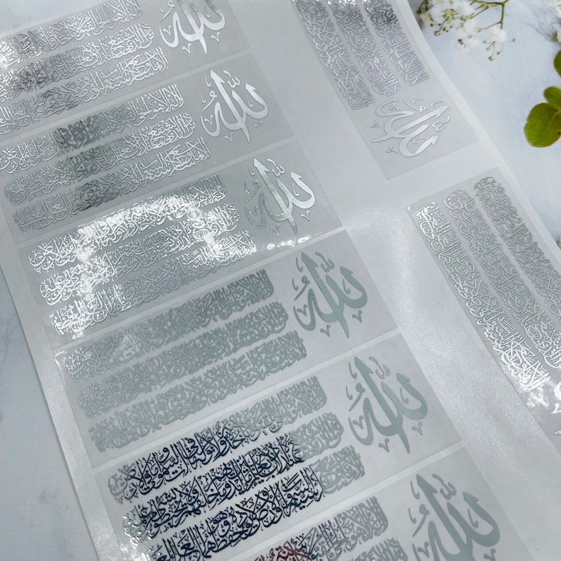 May include: A sheet of adhesive decals featuring silver metallic Arabic calligraphy. The design includes the word Allah in a prominent script, accompanied by intricate, repeating lines of detailed Arabic text arranged in horizontal rows.