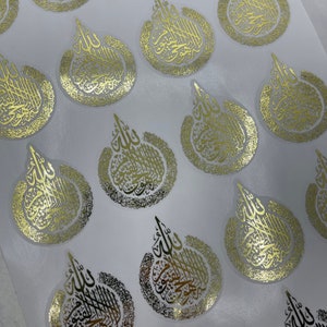 May include: Gold foil stickers with Arabic calligraphy in a teardrop shape. The stickers have a clear background and are arranged on a white surface. The calligraphy is detailed and ornate, with a metallic sheen.