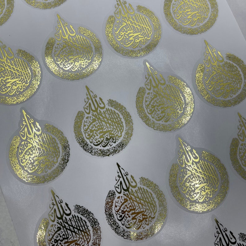 May include: Gold foil stickers with Arabic calligraphy in a teardrop shape. The stickers have a clear background and are arranged on a white surface. The calligraphy is detailed and ornate, with a metallic sheen.