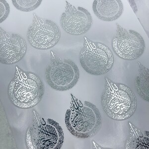 May include: Silver foil stickers with Arabic calligraphy in a teardrop shape. The stickers are arranged on a white background, with the calligraphy in a reflective, metallic finish. The design is intricate and detailed.