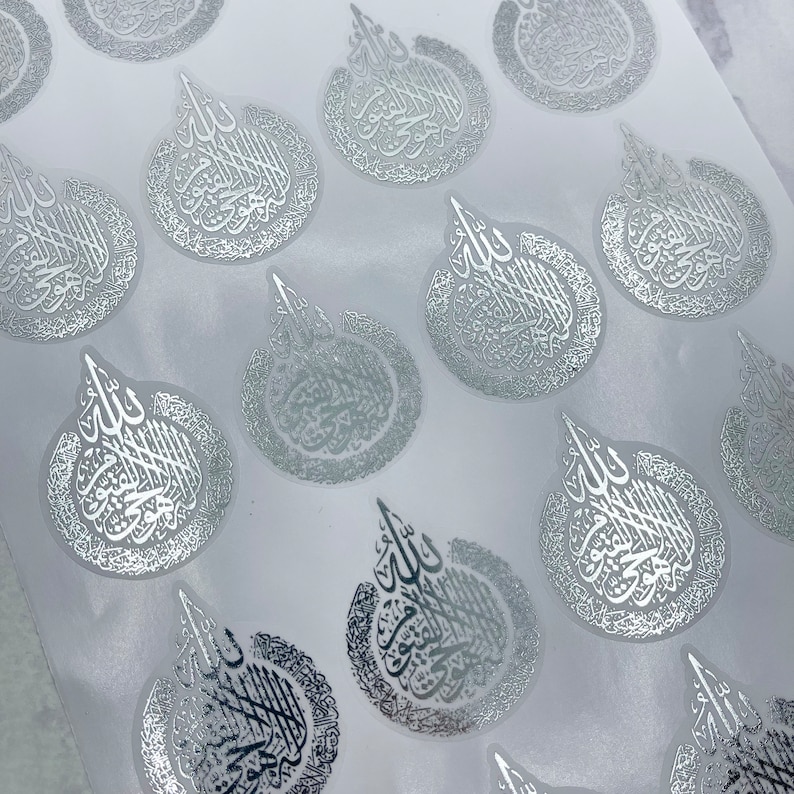 May include: Silver foil stickers with Arabic calligraphy in a teardrop shape. The stickers are arranged on a white background, with the calligraphy in a reflective, metallic finish. The design is intricate and detailed.