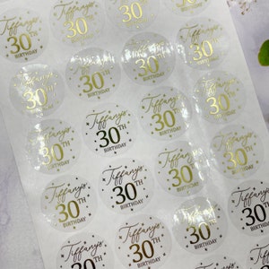 May include: A sheet of clear, round stickers with gold foil lettering. Each sticker says "Tiffany's 30th Birthday" with a gold star design.