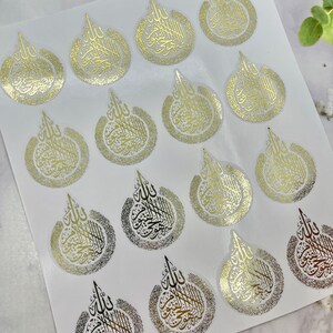 May include: Sheet of gold foil stickers with Arabic calligraphy in a teardrop design. The stickers have a metallic sheen and are arranged on a white background. The calligraphy is in a stylized script.