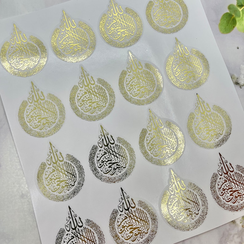 May include: Sheet of gold foil stickers with Arabic calligraphy in a teardrop design. The stickers have a metallic sheen and are arranged on a white background. The calligraphy is in a stylized script.