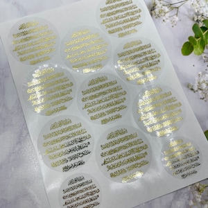 May include: A sheet of clear stickers with gold Arabic calligraphy. The stickers are round and have a white background. The calligraphy is in a traditional style and is very detailed.