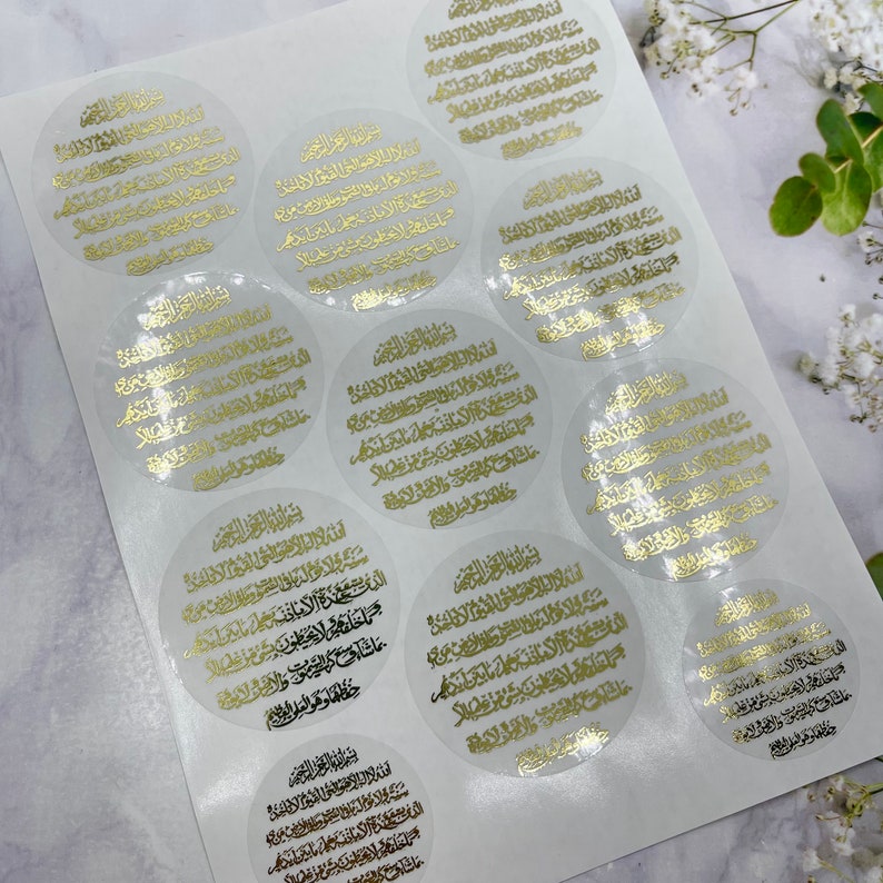 May include: A sheet of clear stickers with gold Arabic calligraphy. The stickers are round and have a white background. The calligraphy is in a traditional style and is very detailed.