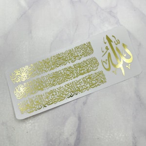 May include: A rectangular white sticker featuring intricate gold foil Arabic calligraphy. The design includes a large central symbol on the right side, with dense, flowing script covering the remaining surface area of the white background.