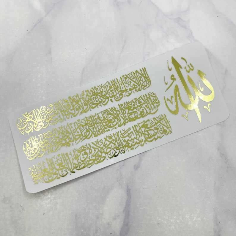 May include: A rectangular white sticker featuring intricate gold foil Arabic calligraphy. The design includes a large central symbol on the right side, with dense, flowing script covering the remaining surface area of the white background.