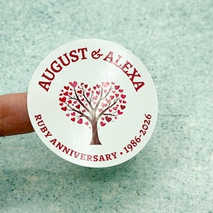 Pode incluir: Um autocolante circular branco com uma árvore vermelho-escura com folhas em forma de coração. O texto AUGUST & ALEXA aparece no topo e RUBY ANNIVERSARY 1986-2026 na parte inferior em letras vermelho-escuras, centrado no gráfico.
