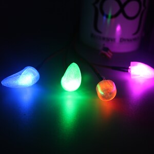 Pu&ograve; includere: Quattro luci LED luminose e colorate in blu, verde, arancione e rosa. Ogni luce ha una forma unica ed &egrave; collegata a un filo sottile. Le luci sono su una superficie scura, con un oggetto bianco sfocato sullo sfondo.