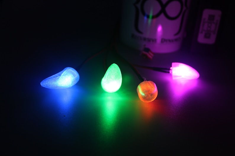 Pu&ograve; includere: Quattro luci LED luminose e colorate in blu, verde, arancione e rosa. Ogni luce ha una forma unica ed &egrave; collegata a un filo sottile. Le luci sono su una superficie scura, con un oggetto bianco sfocato sullo sfondo.