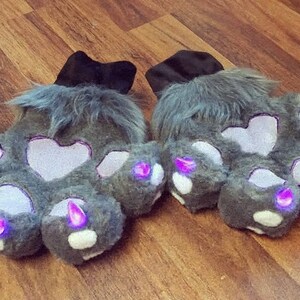 Pu&ograve; includere: Un paio di guanti a zampa grigi e pelosi con cuscinetti a forma di cuore e luci viola brillanti sulle punte delle dita. I guanti hanno polsini neri e sono esposti su una superficie di legno.