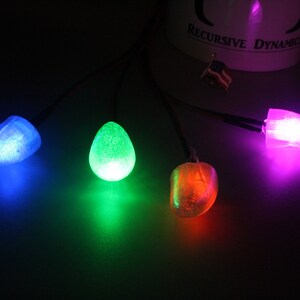 Pu&ograve; includere: Quattro luci LED luminose e colorate in blu, verde, arancione e viola. Ogni luce ha una forma unica, tra cui una goccia e una forma arrotondata. Le luci sono collegate da sottili fili neri, creando un display vibrante su uno sfondo scuro.