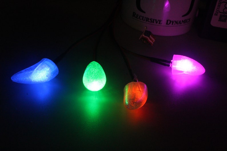 Pu&ograve; includere: Quattro luci LED luminose e colorate in blu, verde, arancione e viola. Ogni luce ha una forma unica, tra cui una goccia e una forma arrotondata. Le luci sono collegate da sottili fili neri, creando un display vibrante su uno sfondo scuro.