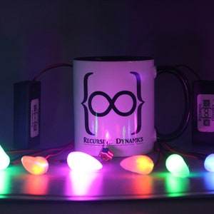 Pu&ograve; includere: Una tazza bianca con bordo e manico neri, con un simbolo dell'infinito nero e la scritta "Recursive Dynamics". La tazza &egrave; illuminata da luci a LED colorate, con piccole luci a forma di uovo che brillano in primo piano.