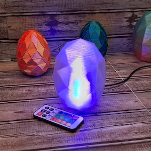 May include: A collection of geometric egg-shaped lamps in various colors, including teal, orange, and purple, are displayed on a wooden surface. A remote control with multiple color options sits in front of the central lamp, which is illuminated with a bright blue light.
