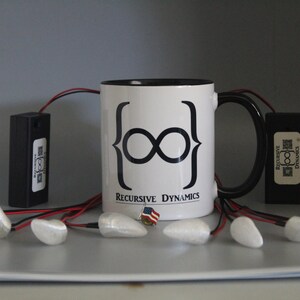 Pu&ograve; includere: Una tazza da caff&egrave; bianca con manico e bordo neri, con un simbolo di infinito nero e la scritta "Recursive Dynamics". L'immagine mostra anche oggetti rettangolari neri, oggetti bianchi a forma di goccia e una piccola spilla con la bandiera americana.