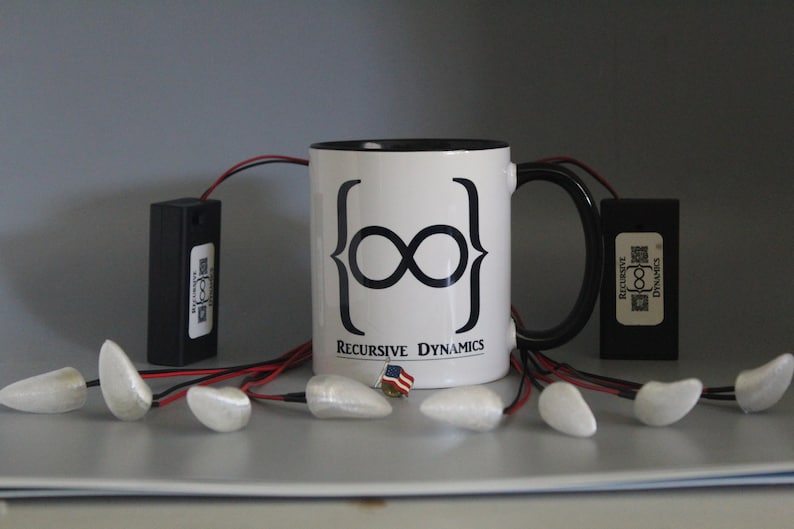 Pu&ograve; includere: Una tazza da caff&egrave; bianca con manico e bordo neri, con un simbolo di infinito nero e la scritta "Recursive Dynamics". L'immagine mostra anche oggetti rettangolari neri, oggetti bianchi a forma di goccia e una piccola spilla con la bandiera americana.