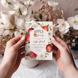 Może przedstawiać: Biała kartka z czerwonymi jabłkami i białymi kwiatami. Tekst na kartce brzmi "Apple of our eye" i "Kaylin Russel". Kartka jest zaproszeniem na baby shower.
