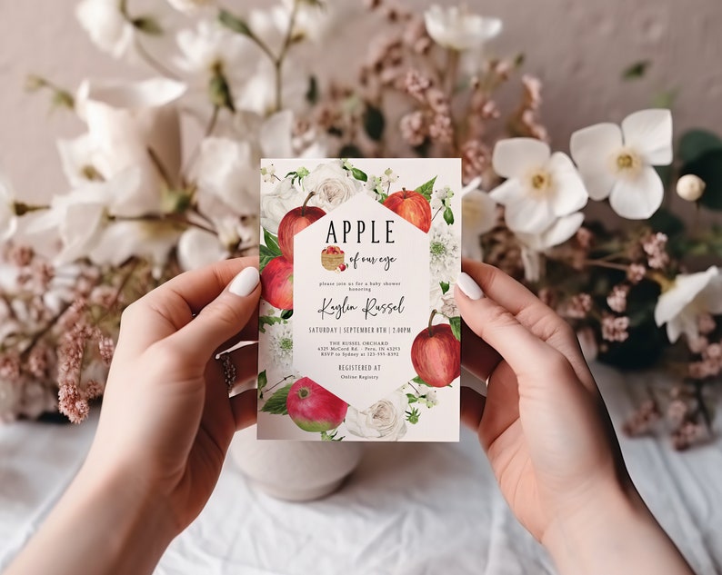 Może przedstawiać: Biała kartka z czerwonymi jabłkami i białymi kwiatami. Tekst na kartce brzmi "Apple of our eye" i "Kaylin Russel". Kartka jest zaproszeniem na baby shower.