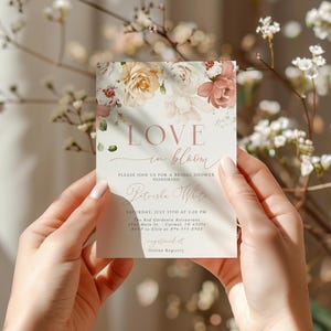 May include: A bridal shower invitation card with floral accents and the words "LOVE in bloom." The invitation features a soft color palette of blush, cream, and green. The card is held by a person's hands.