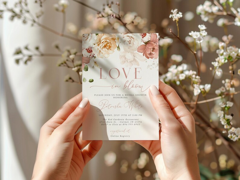 May include: A bridal shower invitation card with floral accents and the words "LOVE in bloom." The invitation features a soft color palette of blush, cream, and green. The card is held by a person's hands.