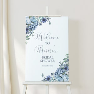 May include: A white welcome sign with blue watercolour flowers and greenery. The text reads "Welcome to Aurora's Bridal Shower September 15th".