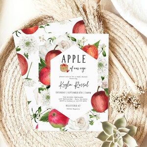 Może przedstawiać: Białe zaproszenie na baby shower z akwarelową ilustracją czerwonych jabłek, białych kwiat&oacute;w i zielonych liści. Tekst brzmi "APPLE of our eye" i "Kaylin Russel".