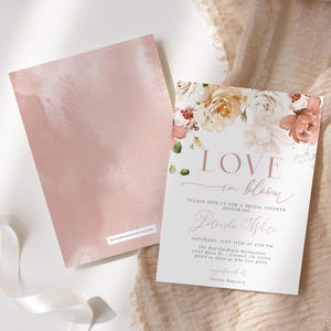May include: Two blush-colored cards, one with a floral design and the text "LOVE in bloom" for a bridal shower. The other card has a watercolor wash. The cards are on a white surface with a ribbon and fabric.