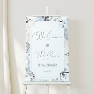 May include: A bridal shower welcome sign with a floral design in white and blue hues. The sign says "Welcome to Millie's Bridal Shower" and the date is January 10th. It is displayed on a white easel, suitable for a wedding.