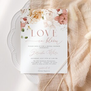 May include: A bridal shower invitation with floral accents. The card features the words "LOVE in bloom" in a delicate script, along with event details. The invitation is set on a white plate and a peach-colored fabric.
