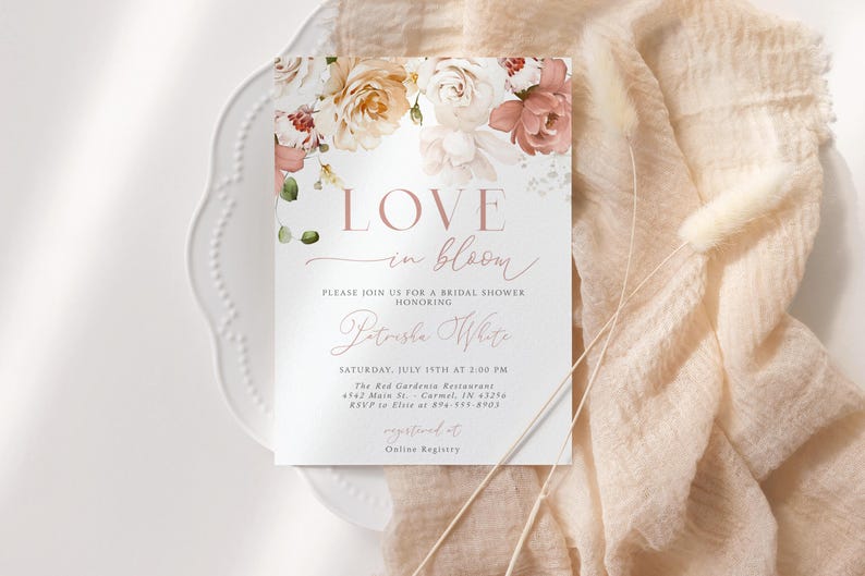 May include: A bridal shower invitation with floral accents. The card features the words "LOVE in bloom" in a delicate script, along with event details. The invitation is set on a white plate and a peach-colored fabric.