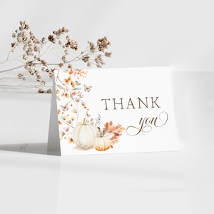 May include: A white folded card with the words "THANK you" in brown script. The card features watercolor illustrations of pumpkins, fall leaves, and butterflies. Dried flowers are in the background.